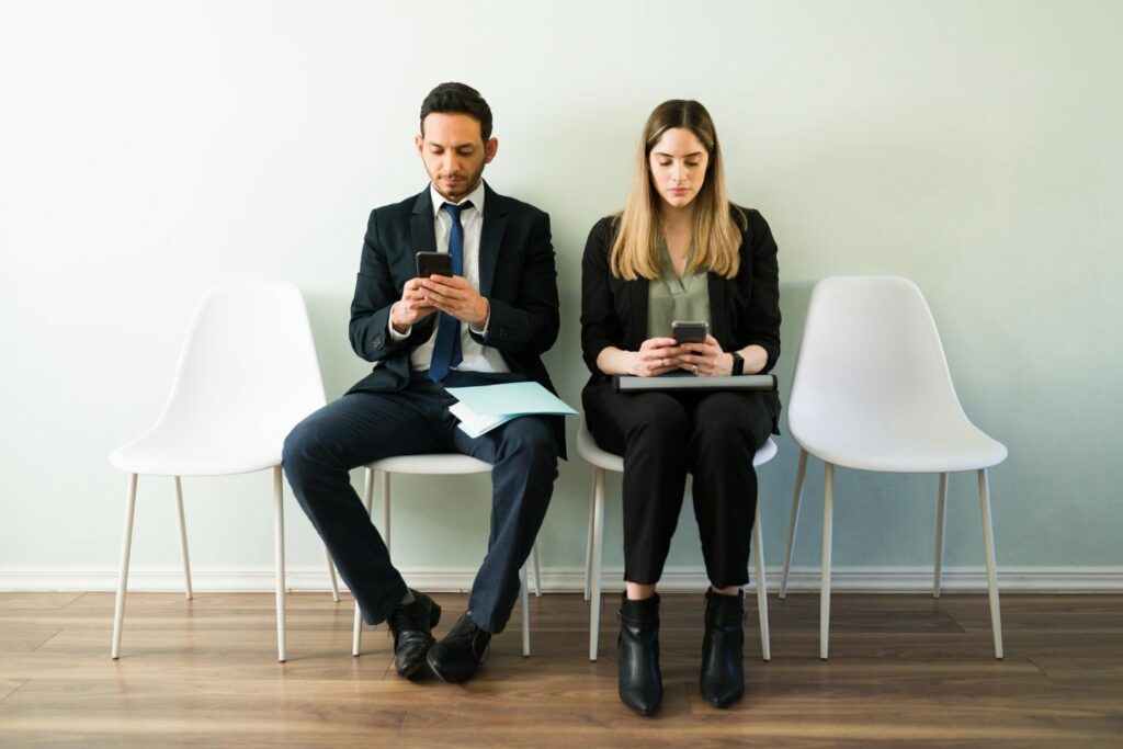 young man and woman waiting sitting next to each other against a wall on white office chairs checking their phones.