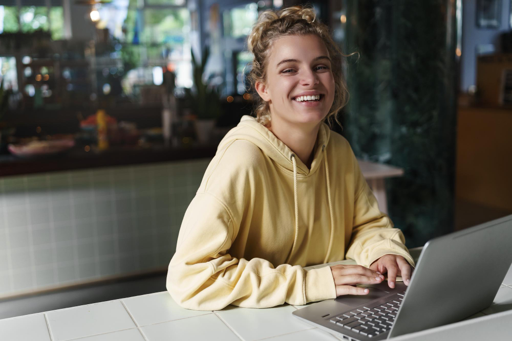 young woman wearing a yellow hoodie sitting in a cafe working on a laptop smiling and looking into the camera.
