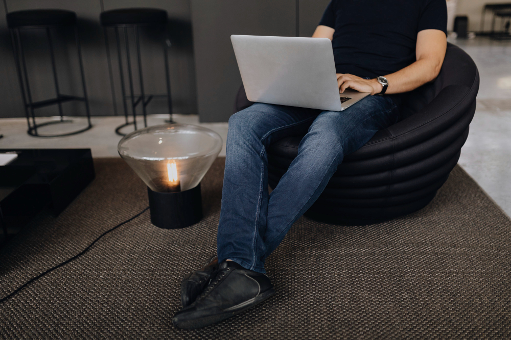 body of a man sitting on a bean bag working on a laptop. He's wearing blue jeans and a black t-shirt and the laptop is grey.